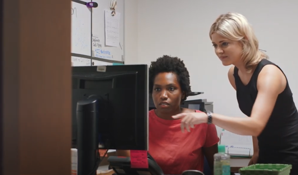 Two women working together at a desk, one seated and looking at a computer monitor while the other stands beside her, pointing at the screen. Office supplies and notes are visible in the background.